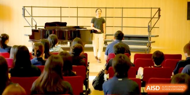An instructor stands on a stage addressing an audience seated in a theater at Beijing Eastbank Academy of Music.