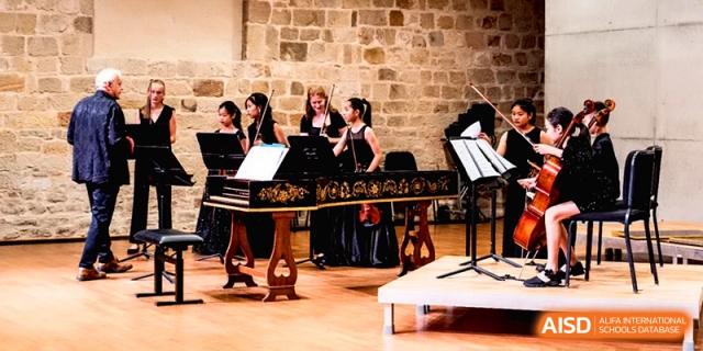 Students performing on stage with a decorated harpsichord and music stands in a stone-walled hall at Beijing Eastbank Academy of Music.