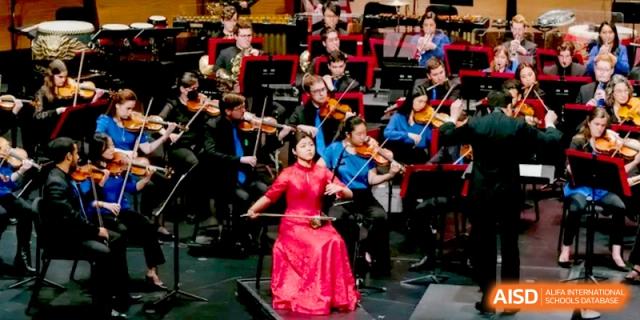 A student in a bright red dress performing as a soloist with a full orchestra at Beijing Eastbank Academy of Music.
