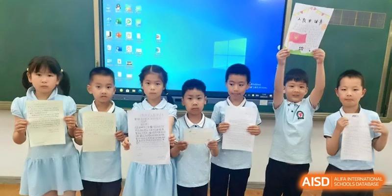 Six elementary school students in uniform stand proudly in a classroom holding up their merit certificates in front of a blue digital screen at Beijing Haidian Rising Experimental School