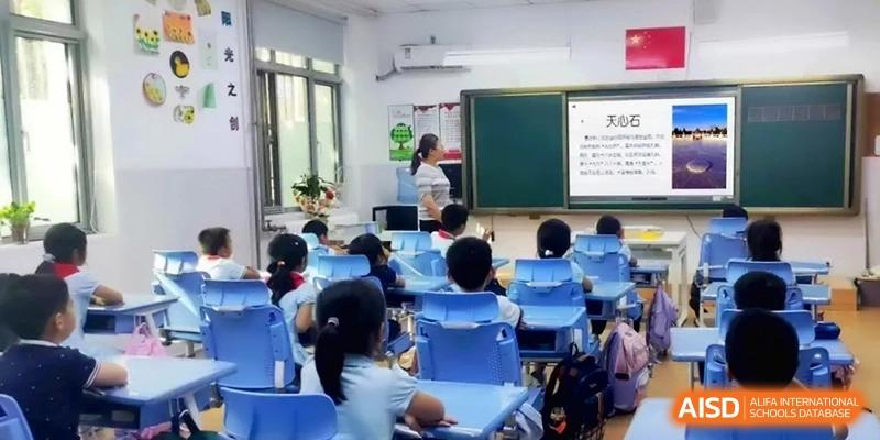 Students sit at their blue desks facing a teacher who is standing by a whiteboard and a large digital display during a primary lesson at Beijing Haidian Rising Experimental School