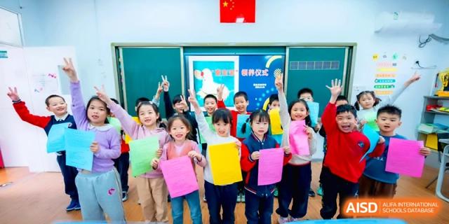 A group of joyful students in a colorful classroom cheer and wave vibrant pieces of construction paper during a school activity at the Beijing Haidian Rising Experimental School