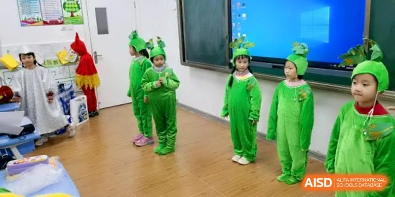 Three young students dressed in bright green frog costumes perform on a stage during a creative arts presentation or school play at the Beijing Haidian Rising Experimental School