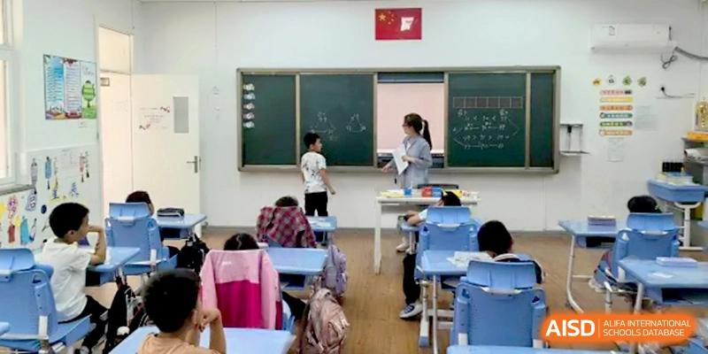 A teacher stands at the front of a modern classroom instructing a young student at the chalkboard while others watch from their desks at Beijing Haidian Rising Experimental School