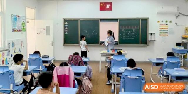 A teacher stands at the front of a modern classroom instructing a young student at the chalkboard while others watch from their desks at Beijing Haidian Rising Experimental School