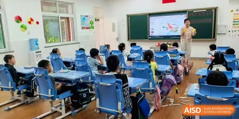 A perspective view of a brightly lit classroom shows primary students sitting at blue desks while a teacher stands by a digital screen at Beijing Haidian Rising Experimental School
