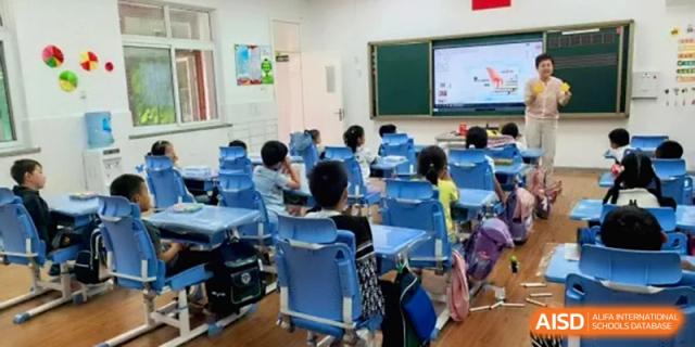 A perspective view of a brightly lit classroom shows primary students sitting at blue desks while a teacher stands by a digital screen at Beijing Haidian Rising Experimental School