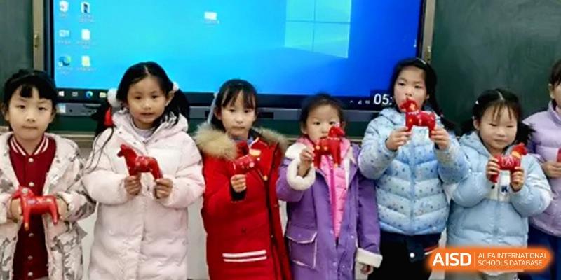 Four young girls wearing warm winter jackets stand together in front of a blue digital display holding small red toy figures at the Beijing Haidian Rising Experimental School