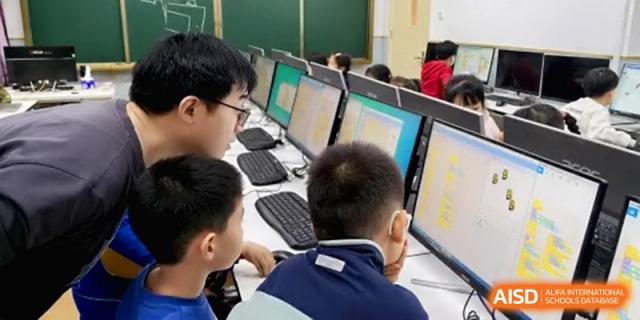 A teacher leans over a row of computer monitors to assist young students during a digital literacy or coding lesson at the Beijing Haidian Rising Experimental School