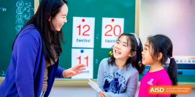 A teacher interacts with a happy young girl in front of a green board displaying numbers twelve and twenty for a math lesson at Beijing Haidian Rising Experimental School