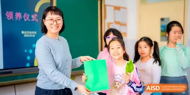 A smiling teacher and student hold up a bright green accordion-style paper craft project together in a classroom setting at the Beijing Haidian Rising Experimental School