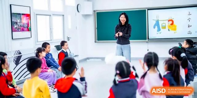 A female teacher stands at the front of a modern classroom gesturing during a lesson while students in red uniforms listen at the Beijing Haidian Rising Experimental School