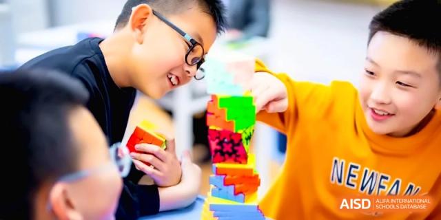 A close-up shot shows a focused student carefully building a tall and colorful tower out of small plastic blocks in class at the Beijing Haidian Rising Experimental School
