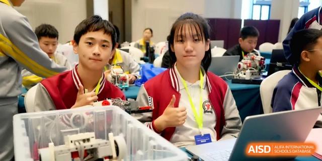 Two older students in red and white tracksuits sit at a desk with technical equipment and give a thumbs up to the camera at the Beijing Haidian Rising Experimental School