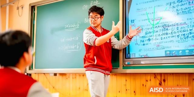 A male teacher in a red vest stands by a digital whiteboard and chalkboard while explaining a lesson to his class at the Beijing Haidian Rising Experimental School