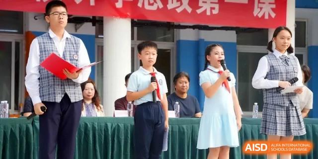 Two students stand at a podium with microphones, leading a school ceremony or presentation on an outdoor stage at the Beijing Haidian Rising Experimental School