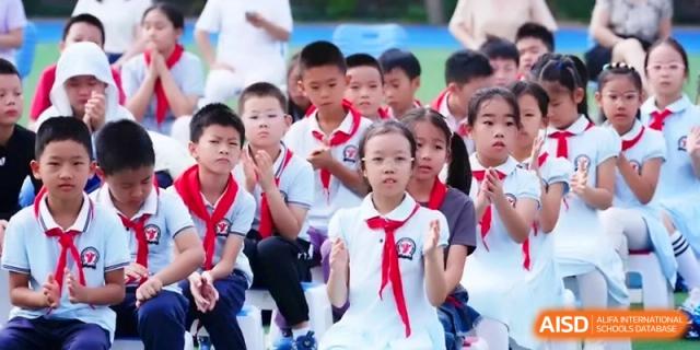 A group of students in uniform with red scarves sit attentively in rows during an outdoor event or presentation at the Beijing Haidian Rising Experimental School