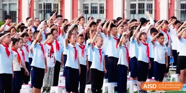 A large group of students stand together on a sports field and perform a synchronized salute during a ceremony at the Beijing Haidian Rising Experimental School