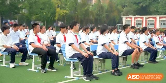 Students in white shirts and red scarves sit in organized rows on a green sports field during a formal school gathering at Beijing Haidian Rising Experimental School
