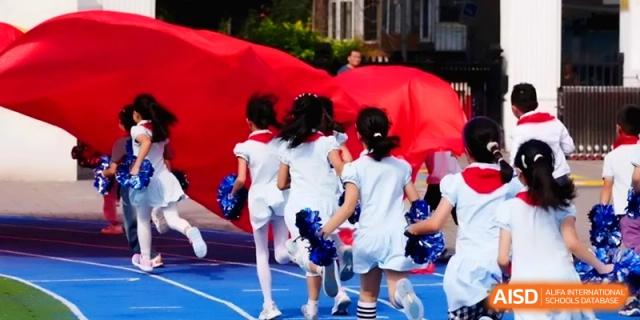 Students in white uniforms run across a blue athletic track while carrying a large red banner during a sports event at Beijing Haidian Rising Experimental School