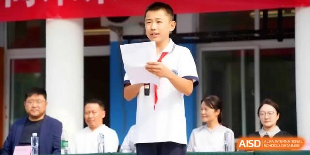 A male student stands outdoors holding a script and speaking into a microphone during a school presentation at the Beijing Haidian Rising Experimental School