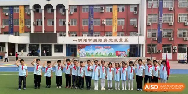 A large group of students in uniform stand in a line in front of a red brick school building for a group photo at Beijing Haidian Rising Experimental School