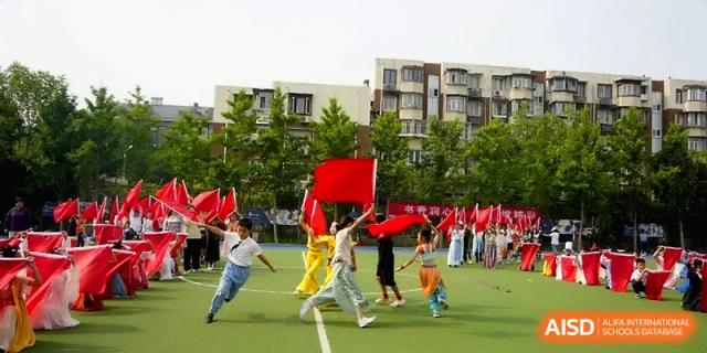 Students run across a green field carrying large red flags during a dynamic outdoor performance or sports event at Beijing Haidian Rising Experimental School