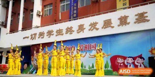 A line of performers in yellow outfits poses during a cultural dance performance in front of a school building at Beijing Haidian Rising Experimental School