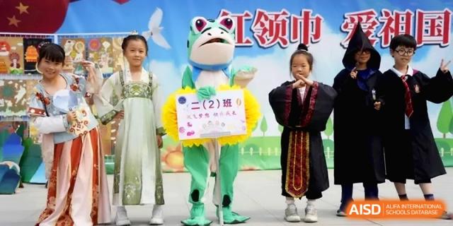 A student in a green frog mascot costume stands on stage with other students in traditional dress at the Beijing Haidian Rising Experimental School