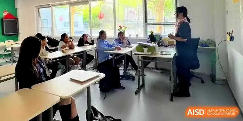 Students sitting at desks in a bright classroom while a teacher leads a discussion near the window at HD Beijing School.