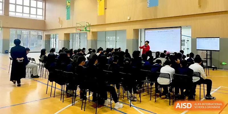 A large assembly of students in dark uniforms sitting in the gymnasium watching a presentation at HD Beijing School.