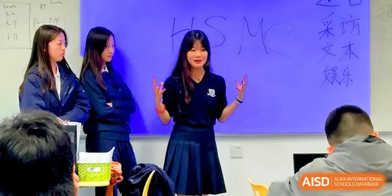 A young female student gesturing while speaking enthusiastically in front of a blue background in a classroom at HD Beijing School.