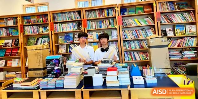 Two students standing behind a table filled with stacks of books in front of a large library bookshelf at HD Beijing School.