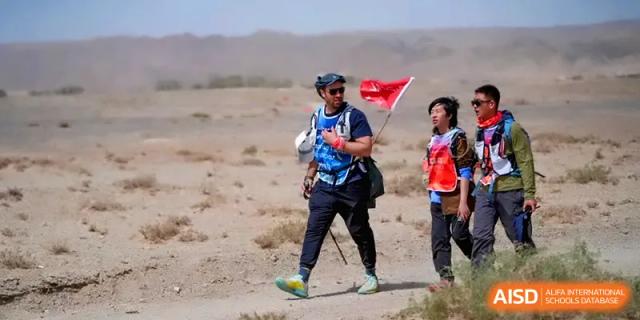 A student hiker wearing a blue vest and carrying a red flag while walking through a desert environment at HD Beijing School.