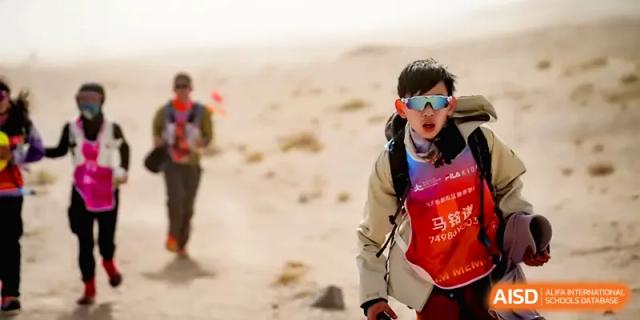 A young hiker wearing glasses and a red safety vest treks through a sandy desert landscape during an outdoor expedition with HD Beijing School.