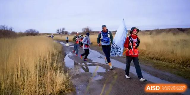 A group of hikers in athletic gear walks along a muddy path through a field during a long-distance trekking event at HD Beijing School.
