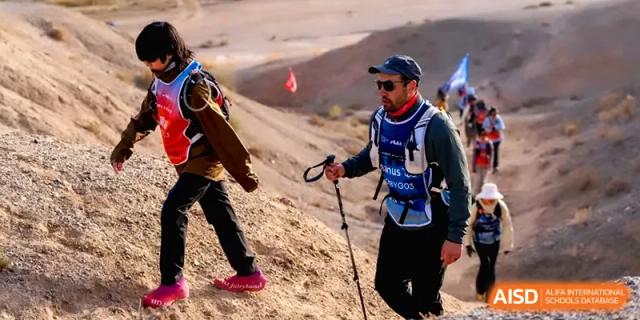 An adult leader with trekking poles guides a group of students up a rugged, sandy hill during a desert hike at HD Beijing School.