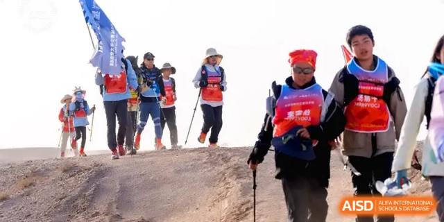 A line of student hikers wearing backpacks and safety vests reaches the crest of a hill during a trekking trip at HD Beijing School.
