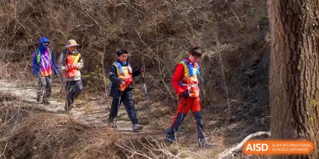 Two students in hiking gear use trekking poles to navigate through a dry, brushy trail during an outdoor challenge at HD Beijing School.