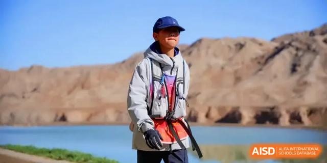 A smiling student in a grey jacket and blue cap stands in front of a mountainous desert backdrop during a trip with HD Beijing School.