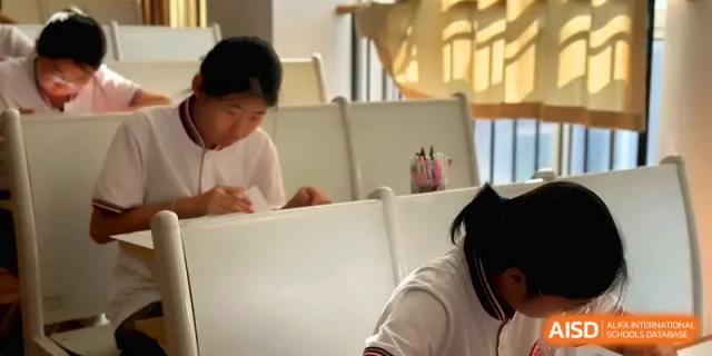 A student in a white uniform works diligently at their desk in a sunny classroom at Qingdao Experimental High School.