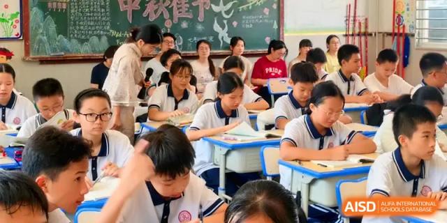 Students in white and blue uniforms sit at their desks in a bright classroom, listening intently during a lecture or presentation at Quanzhou No. 5 Middle School.