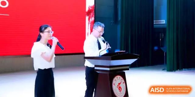 A faculty member speaks into a microphone at a lectern during a formal school celebration or assembly at Quanzhou No. 5 Middle School.