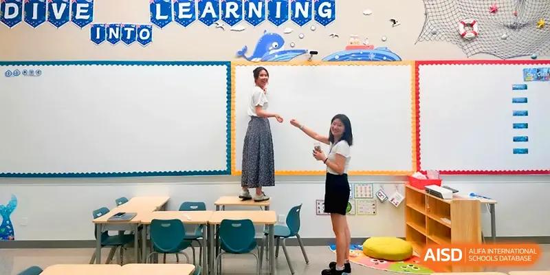Two teachers interact in a bright classroom with blue chairs and educational posters at Shenzhen Mingwan School.