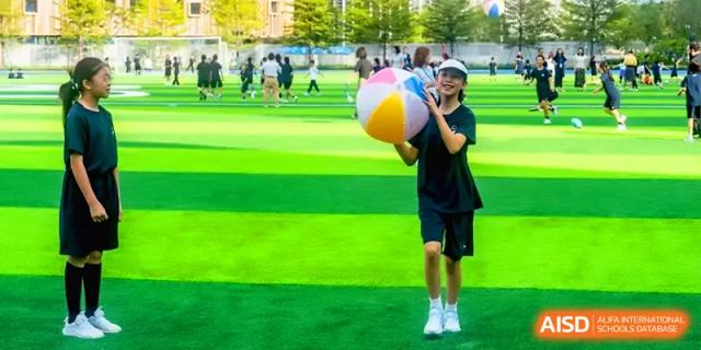 Young student smiling and holding a large, colorful beach ball on a green sports field at Shenzhen Mingwan School.