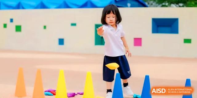 A small child focuses intently on tossing a yellow bean bag over a line of cones at Shenzhen Mingwan School.