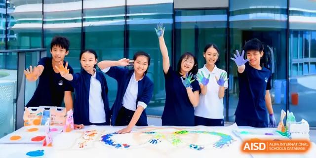 Group of smiling girls in school uniform raising blue-painted hands next to their artwork at Shenzhen Mingwan School.