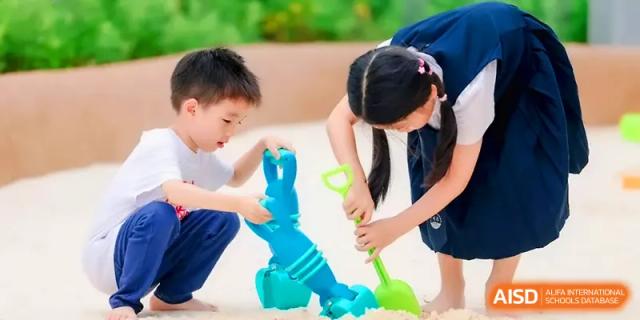 Two students collaborate using blue and green plastic shovels in a sensory play sandbox at Shenzhen Mingwan School.