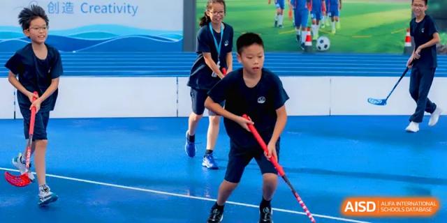 A boy holding a hockey stick and a girl standing behind him on a blue indoor multi-sport court at Shenzhen Mingwan School.