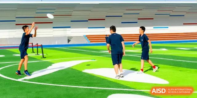 Students in sports kit walking on a green turf field near a grandstand with colorful accents at Shenzhen Mingwan School.